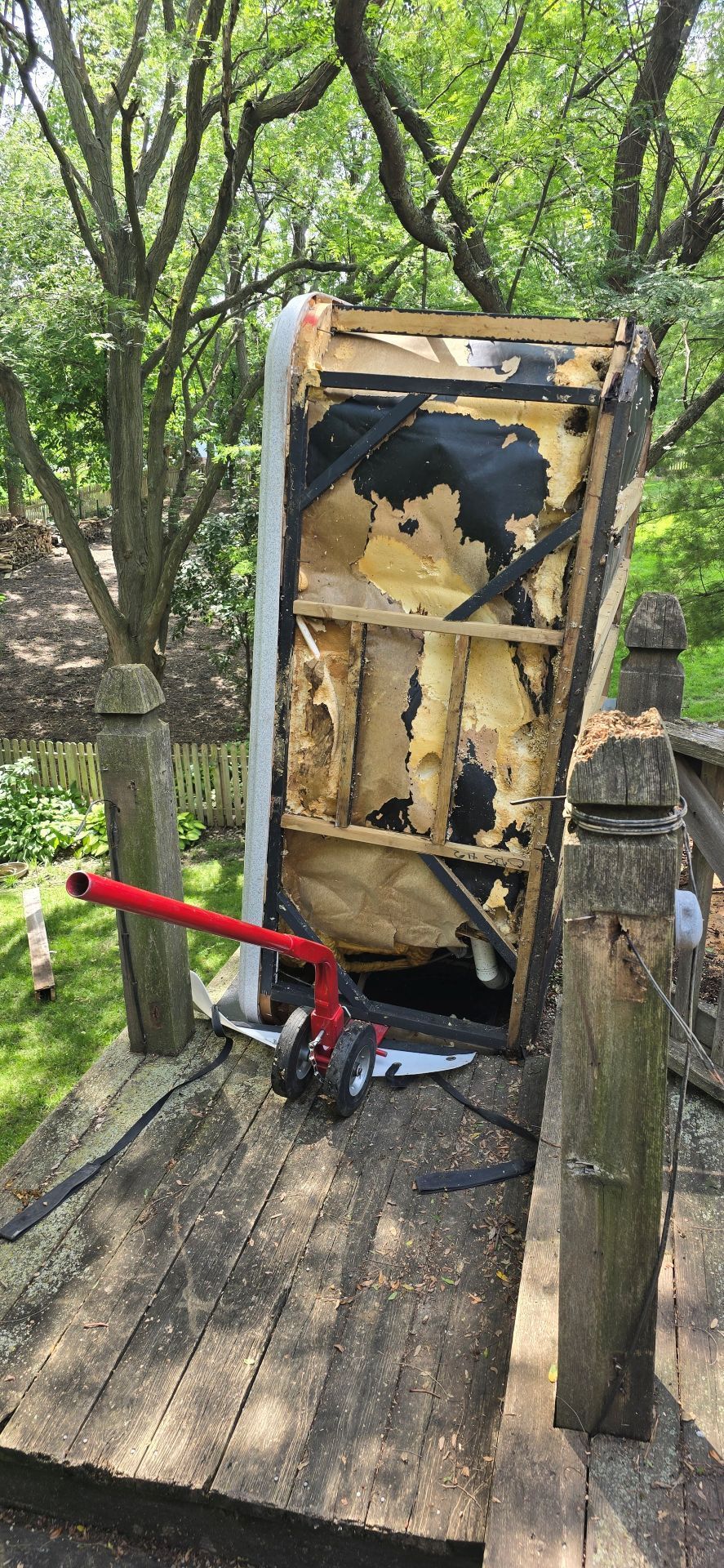 Charred wooden box on a deck with a red-handled tool leaning against it, in a wooded outdoor area