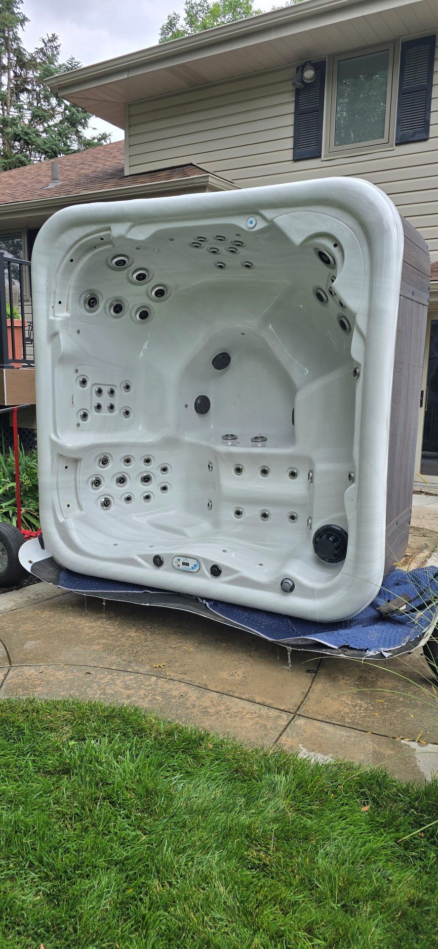 White hot tub shell propped on lawn beside a brick house, with blue tarp underneath.