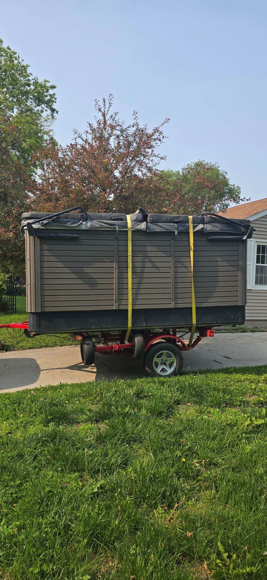 Gray enclosed utility trailer parked in a driveway, strapped with yellow tie-downs in front of a house.