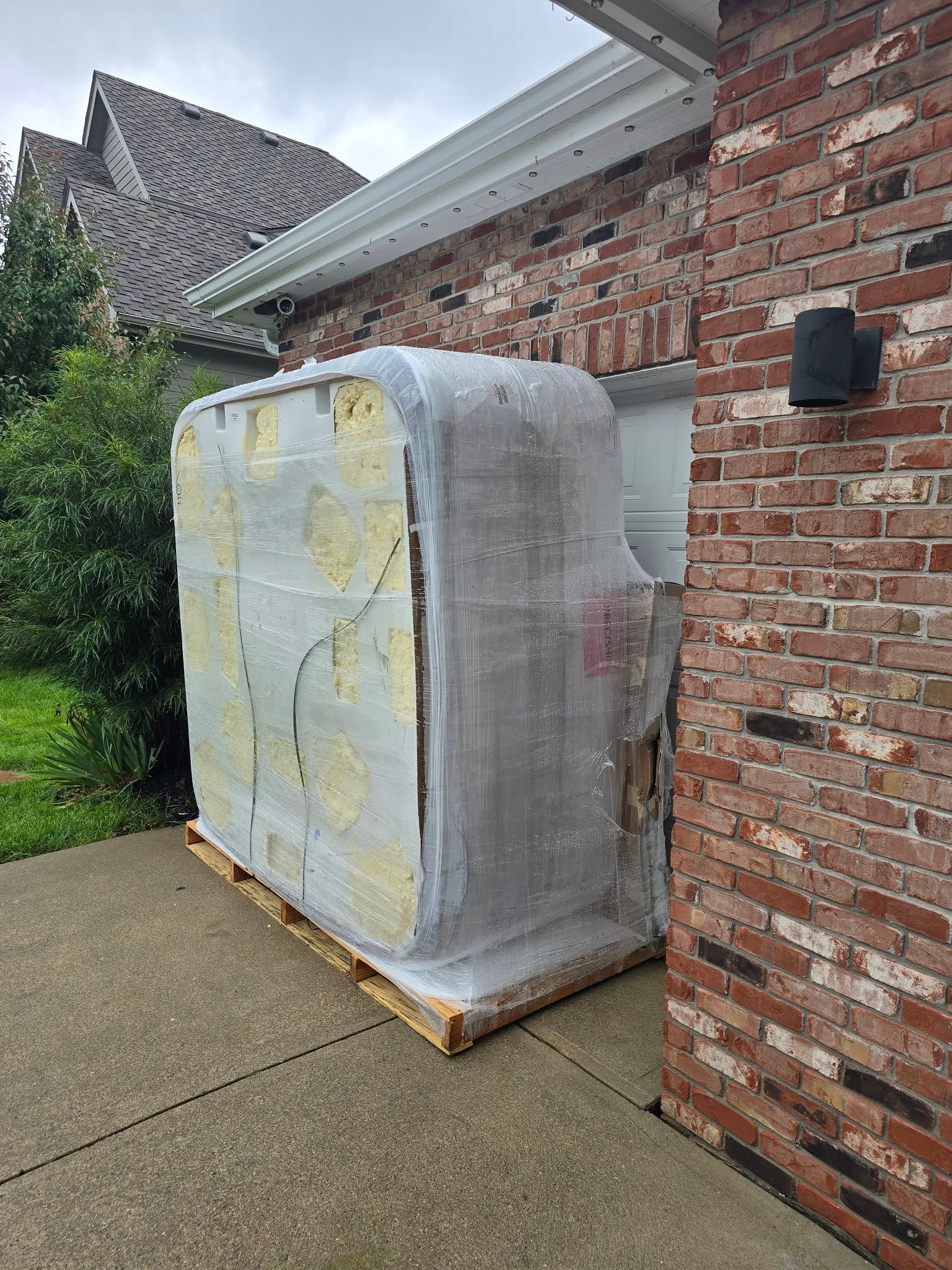 Large shrink-wrapped appliance on a pallet beside a brick house on a concrete driveway.