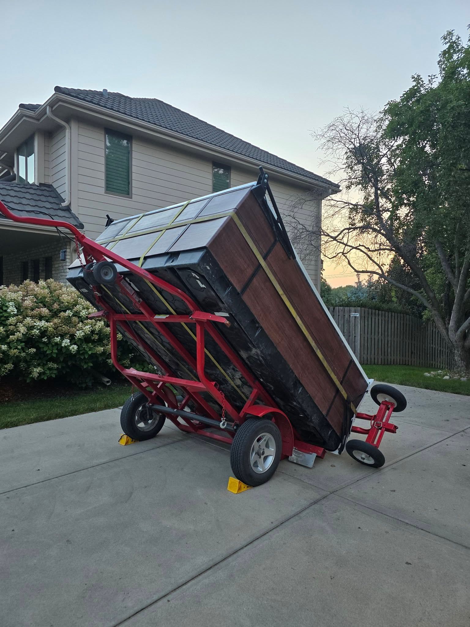 Red hand truck tilting a large wooden shed or panel on a suburban driveway at dusk