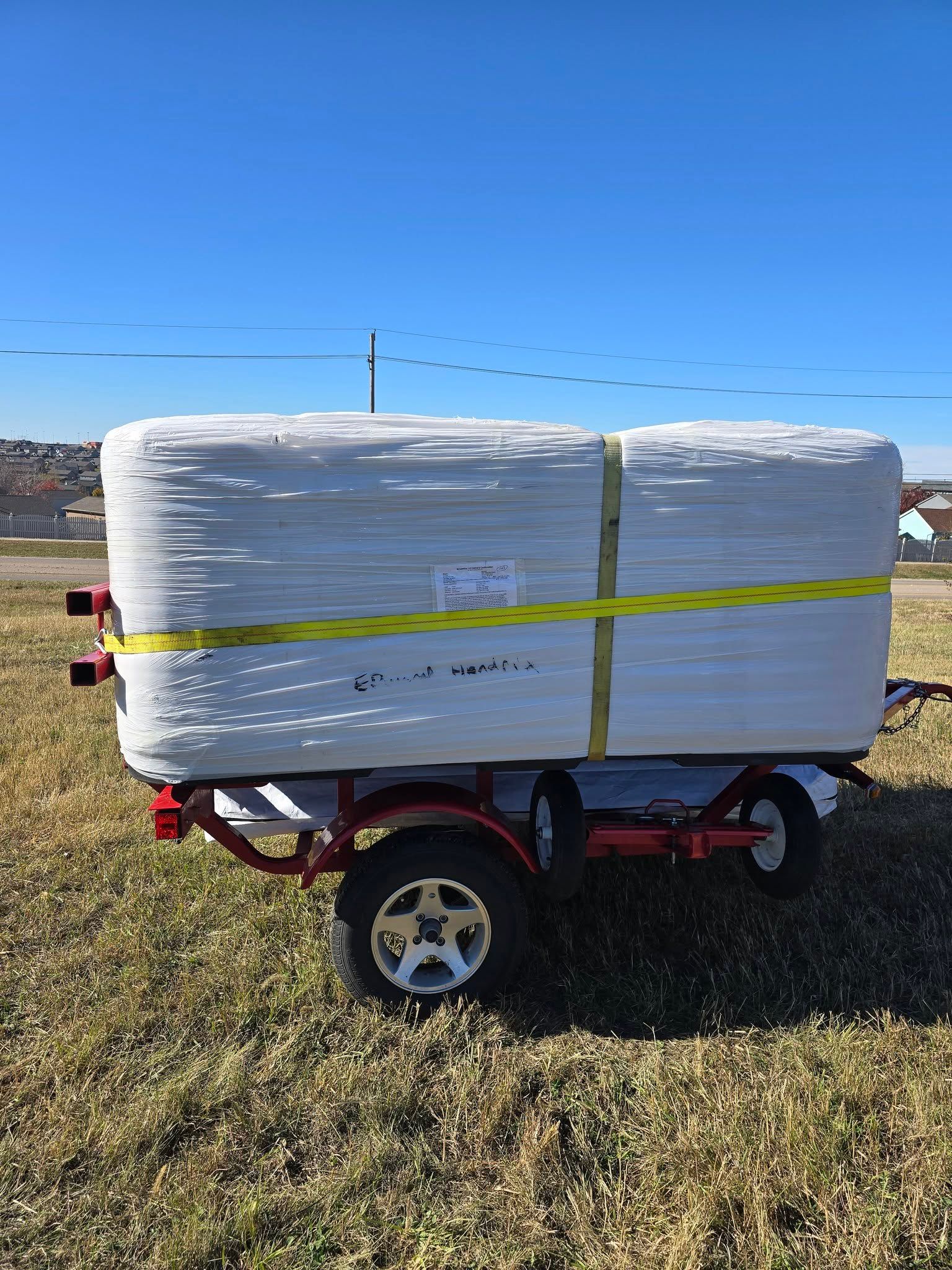 White-wrapped pallet secured with yellow straps on a red trailer in a grassy field under blue sky