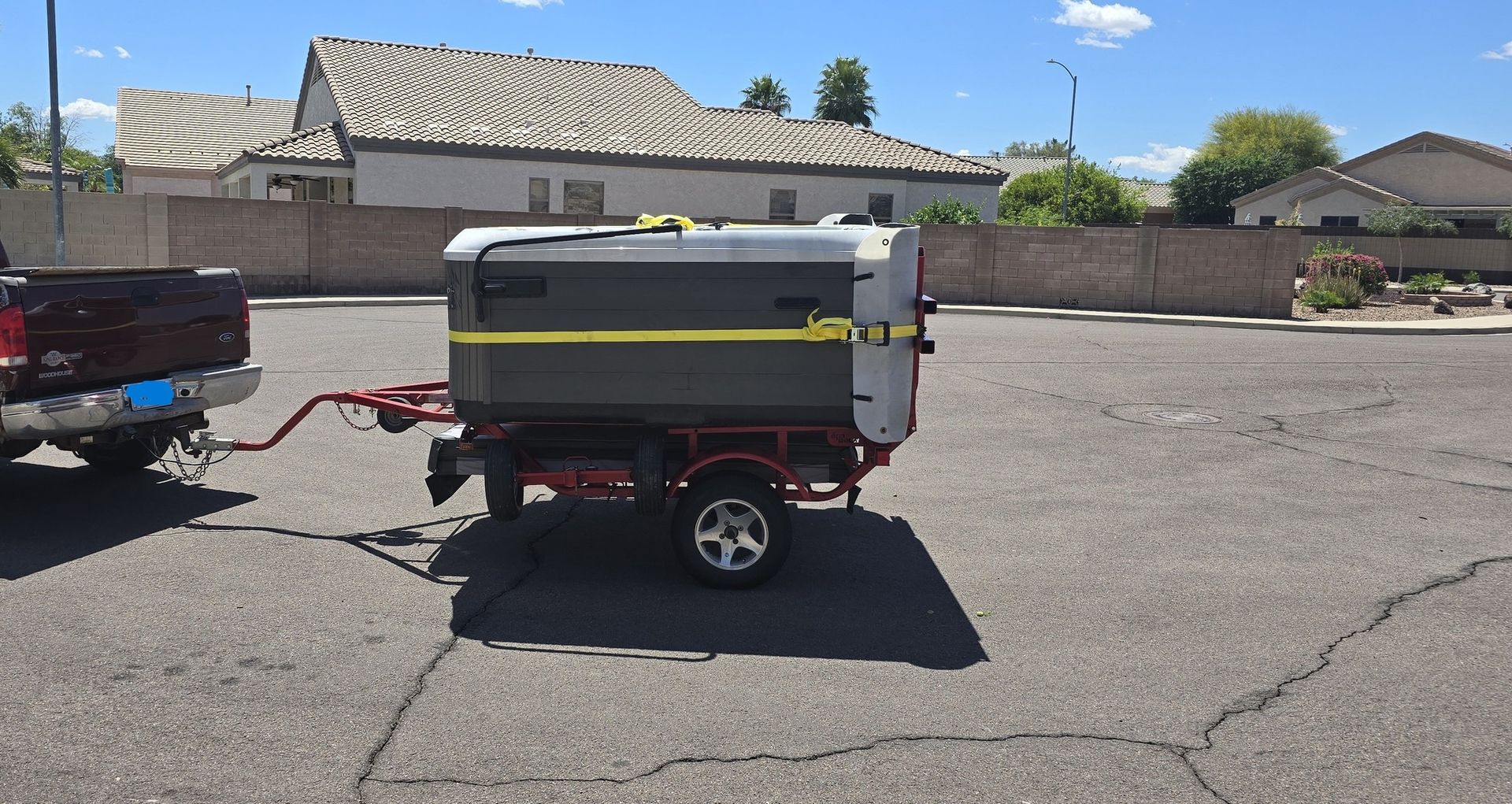 Small gray utility trailer parked in a paved lot, hitched to a red pickup truck.
