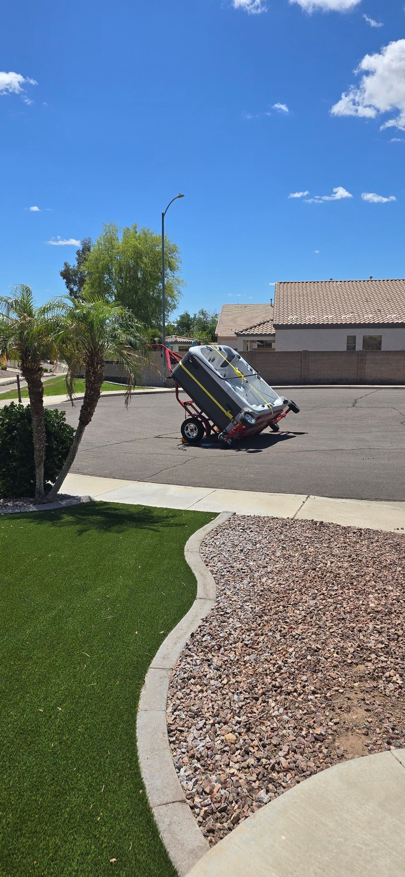 Tipped-over lawn mower on a suburban driveway beside a green lawn and desert landscaping under a blue sky