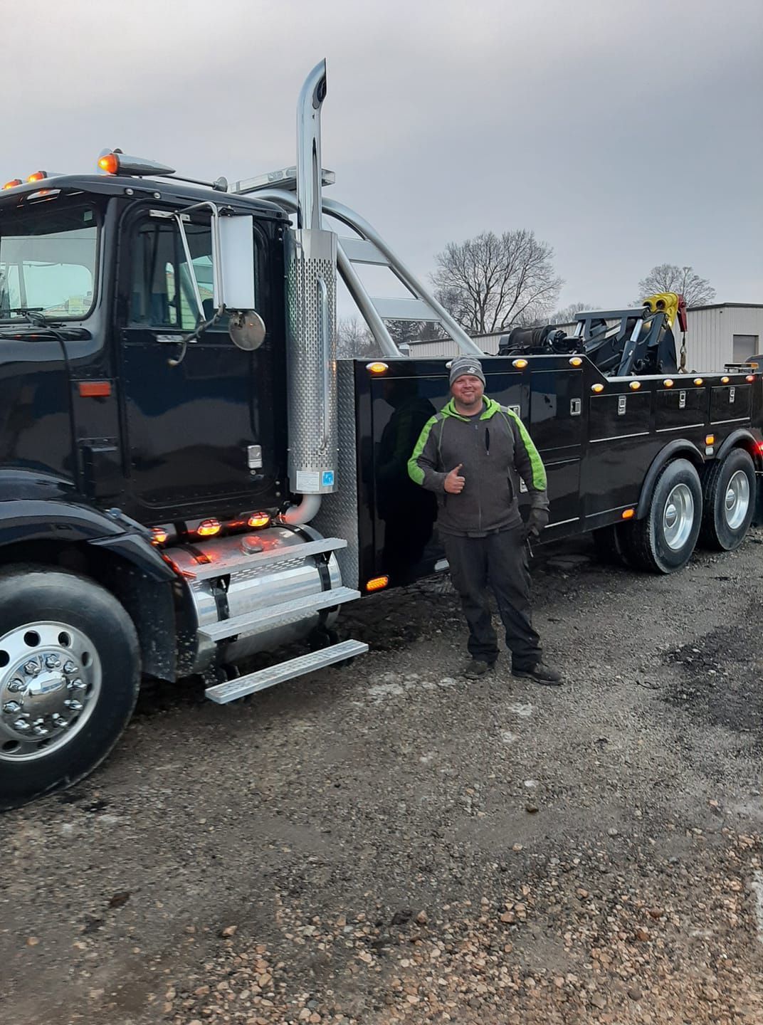 A man is standing in front of a tow truck giving a thumbs up.