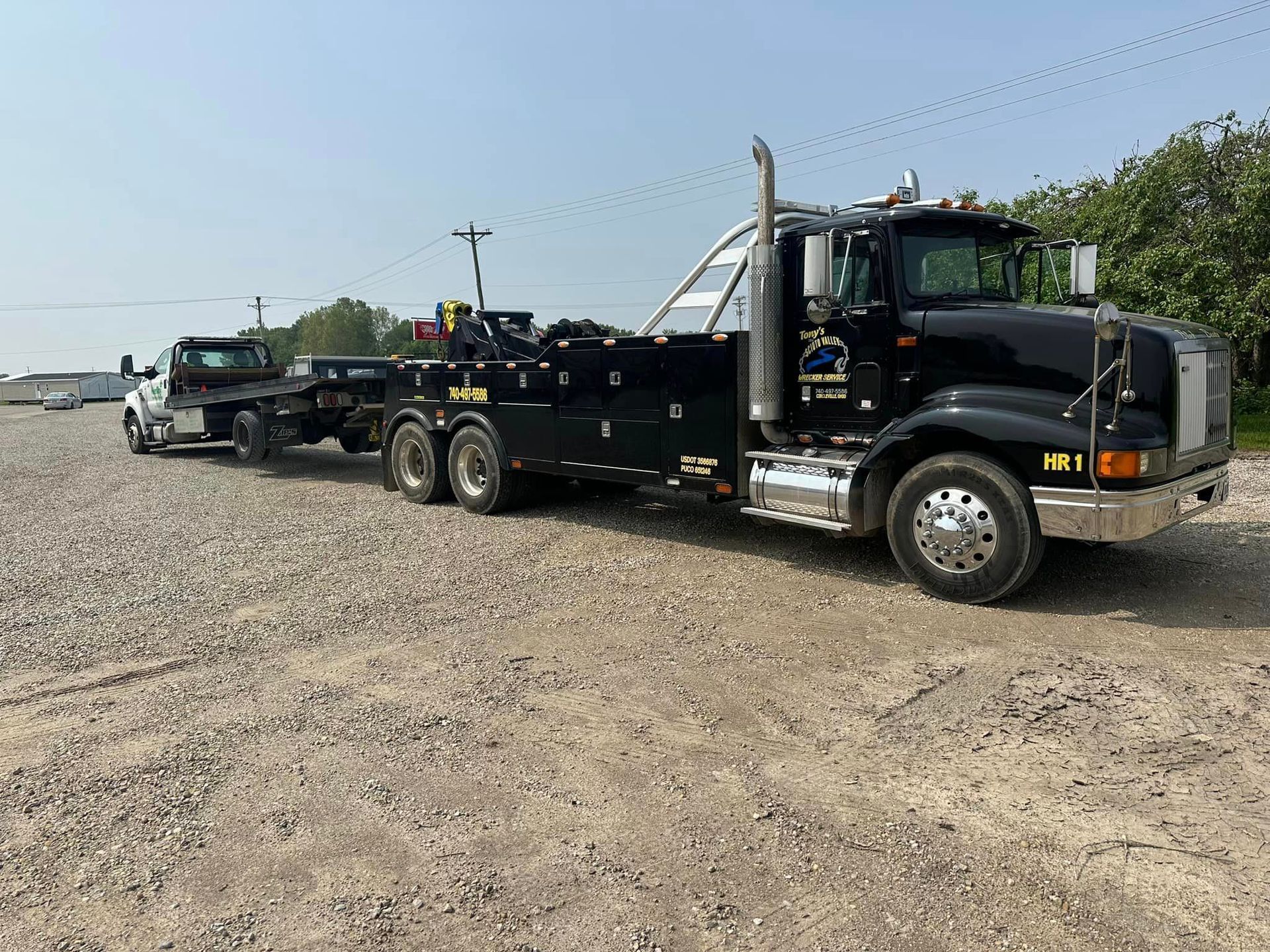 A black tow truck with a trailer attached to it is parked in a gravel lot.