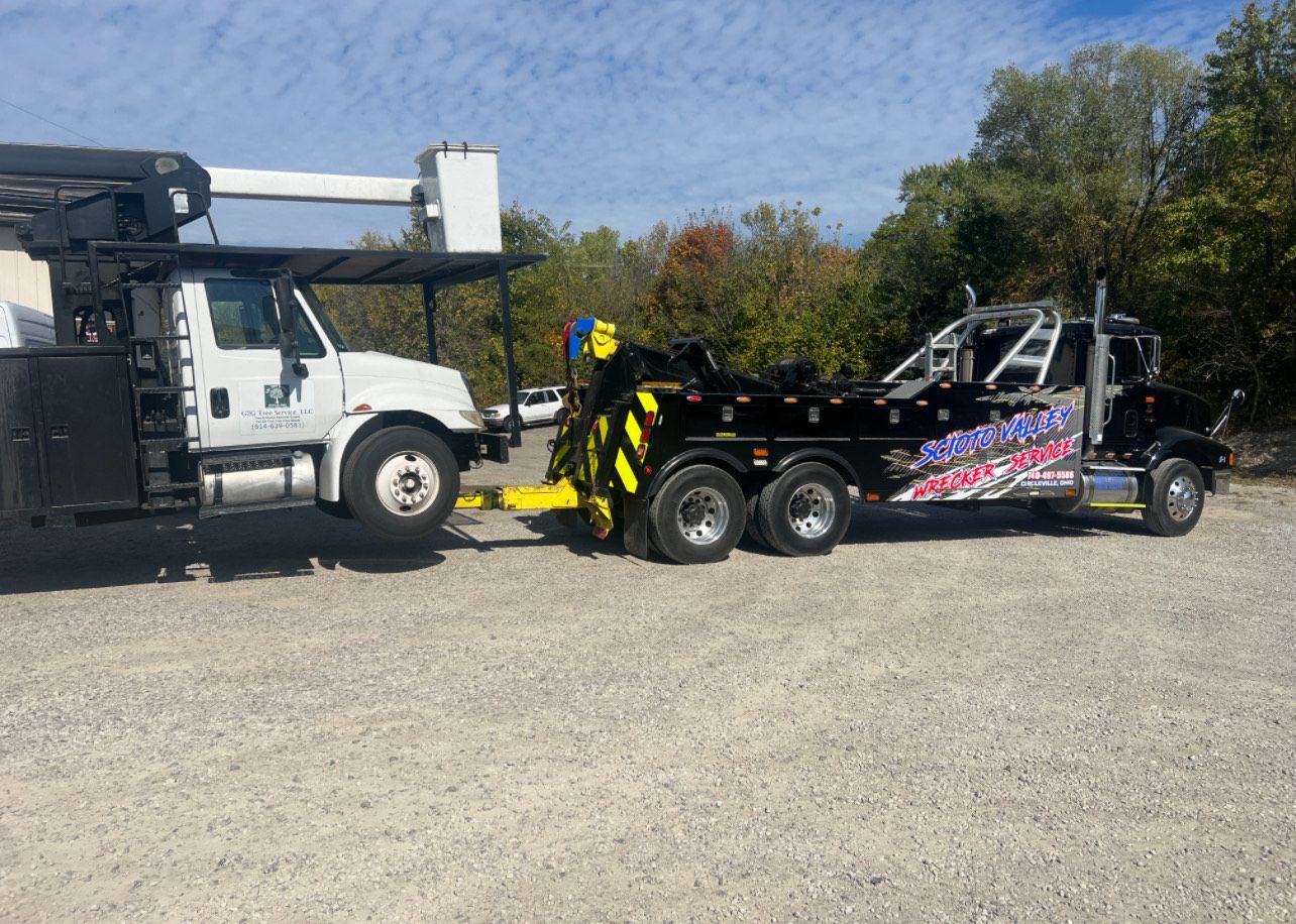 A black tow truck with a trailer attached to it is parked in a gravel lot.
