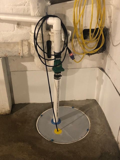 Submersible pump in a concrete basement sump with hoses and coiled cables against a brick wall
