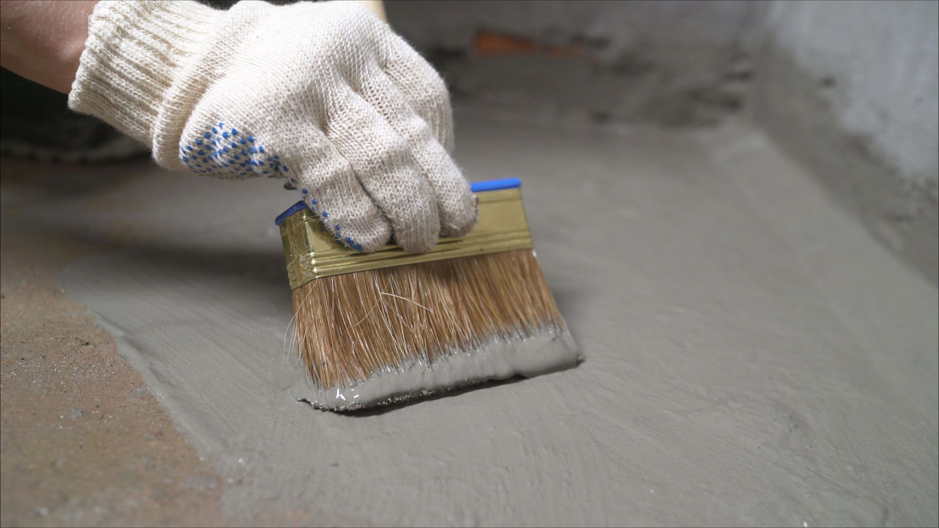 Gloved hand using a brush to spread gray concrete or sealant on a floor surface