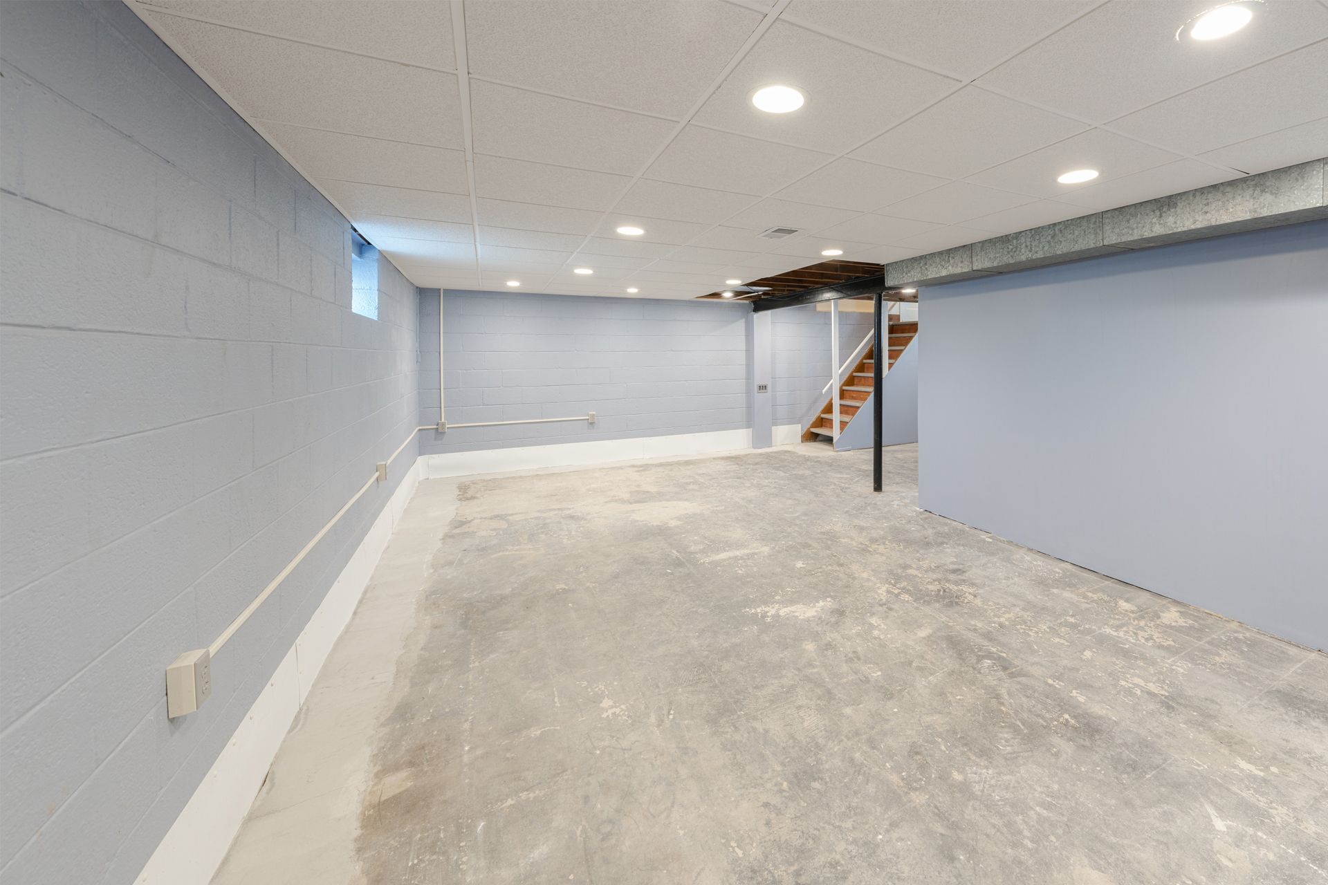 Empty unfinished basement room with gray walls, concrete floor, recessed lights, and stairs at the back.