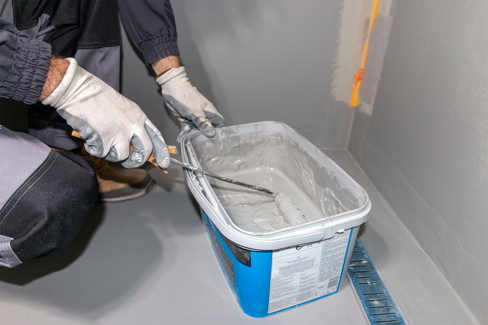 Gloved worker stirring gray coating in a blue bucket on a floor with a trowel, beside a wall and measuring tape.