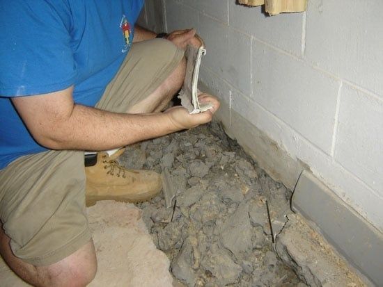 Person in blue shirt applying gray mortar along a basement wall floor joint.