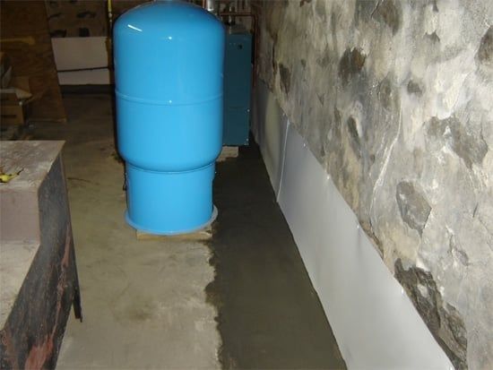 Blue cylindrical tank in a narrow basement corridor beside a rough stone wall.