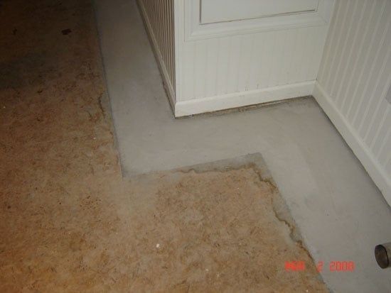 Corner of a room with old carpet removed, exposing gray subfloor near white cabinets and paneling.