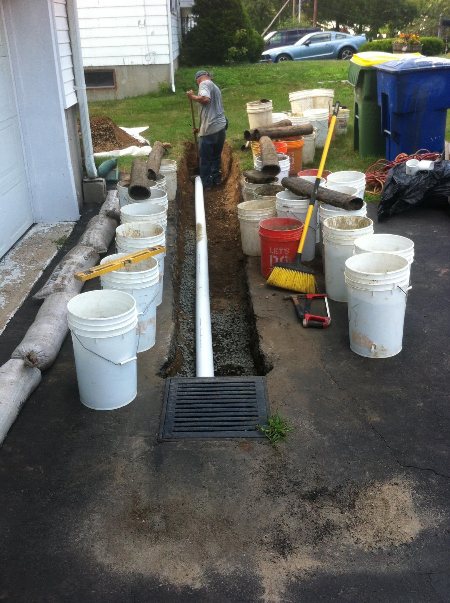 A trench along a driveway under repair, with tools, buckets, and a worker in the background.