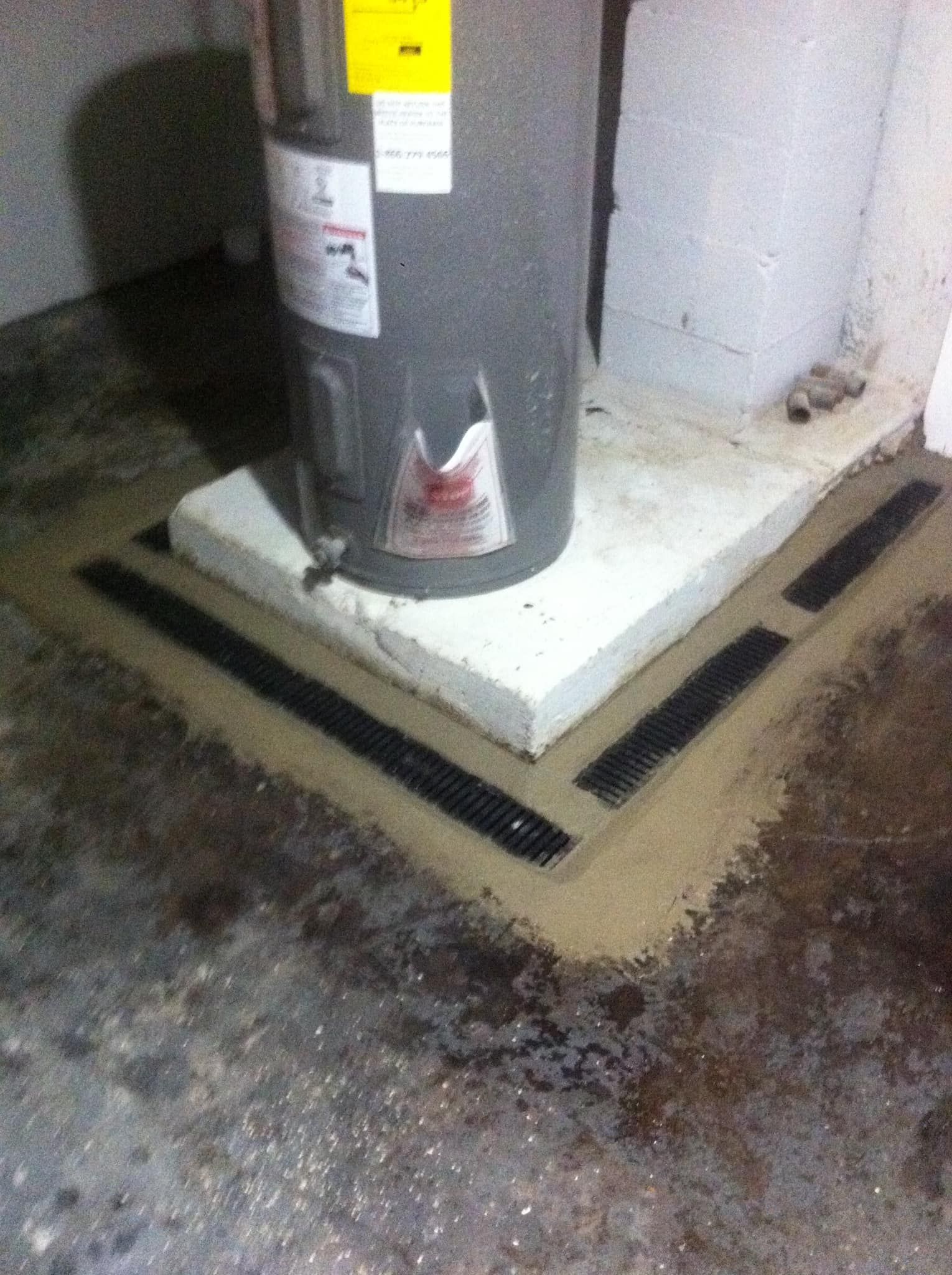 Water heater on a concrete pedestal in a basement corner, with a black moisture or drainage barrier around the base.