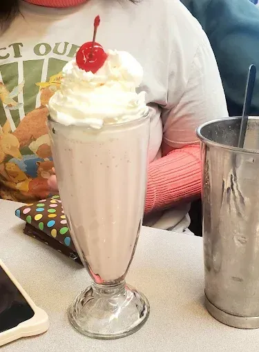 Pink milkshake in a glass with whipped cream and a cherry, next to a metal container.