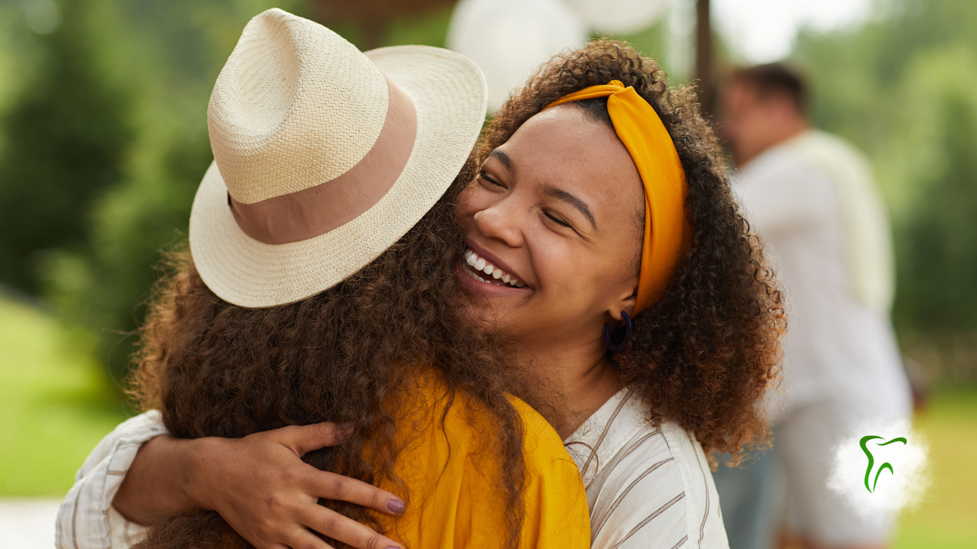 Woman in yellow headband hugs person wearing a straw hat, smiling outdoors.