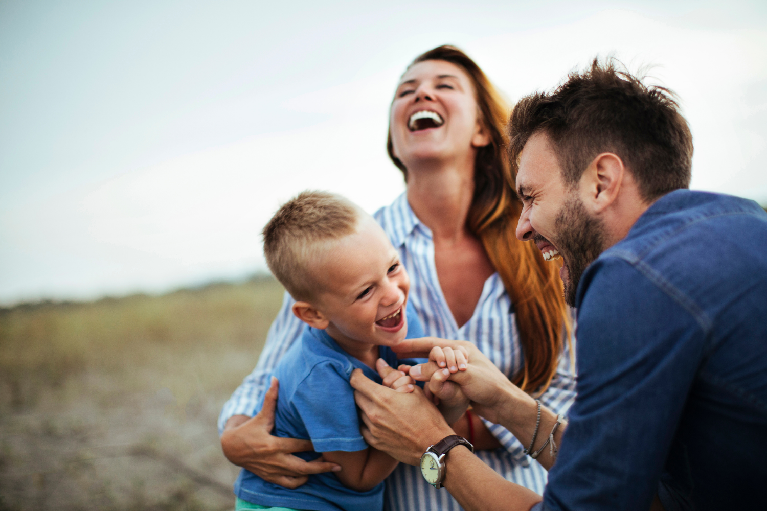 Three people laughing and hugging outdoors in a grassy field, with a child in a blue shirt in the foreground