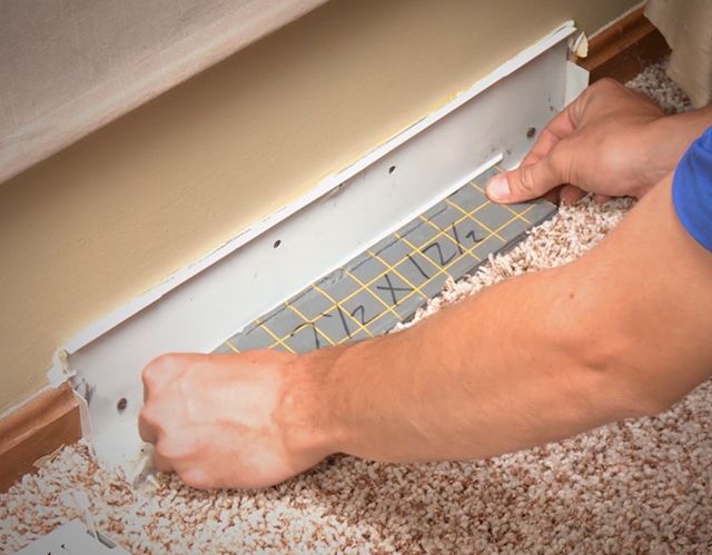 Person installing a vent cover in a wall. The cover is gray, the wall is beige, and the carpet is light brown.