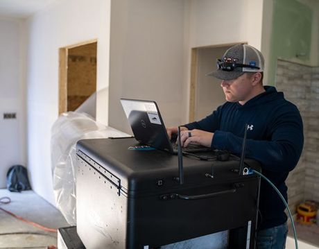 Man in a gray hat and blue hoodie using a laptop on a black box, indoors near construction.