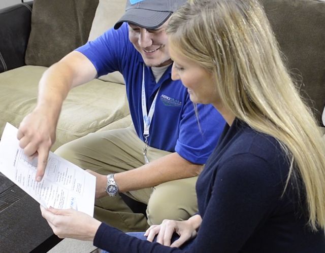 A person in blue shirt points to a document while another person looks on. They sit near a brown couch.