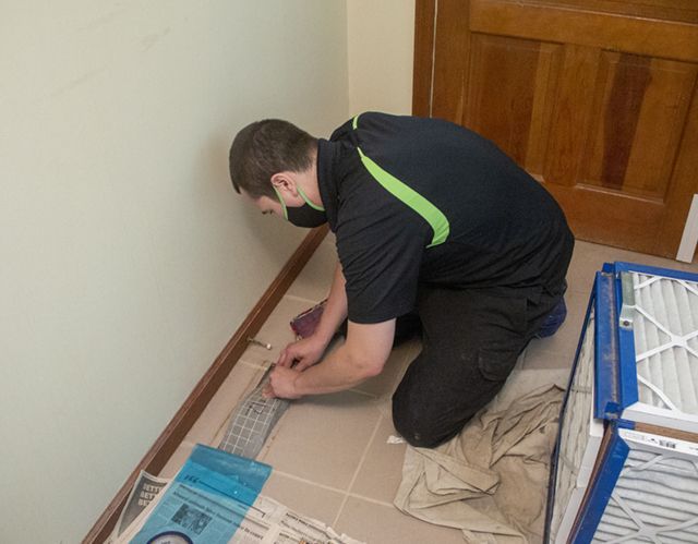 Person kneeling, working on something near a wall. They wear black shirt with neon green and black pants. Filters and newspapers are on the tiled floor.