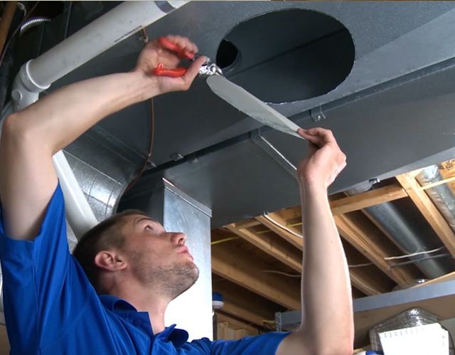 Man in blue shirt working on ductwork, using pliers and tape. Gray ducts in a basement.