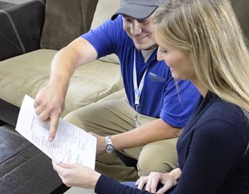 Man in blue shirt points to paperwork while woman smiles, looking at it. They are in a room with a sofa.