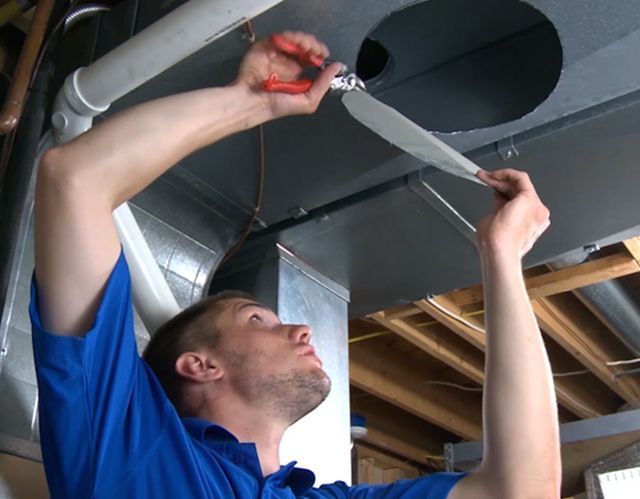 Man in blue shirt working on HVAC ductwork, using pliers.