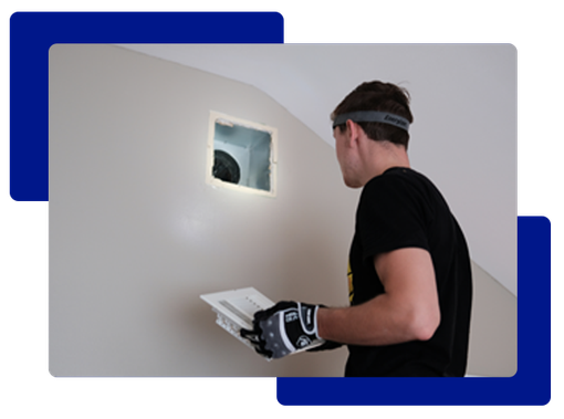 Man inspecting vent, holding paper. Wears safety glasses and gloves. Wall is beige, setting appears to be inside.