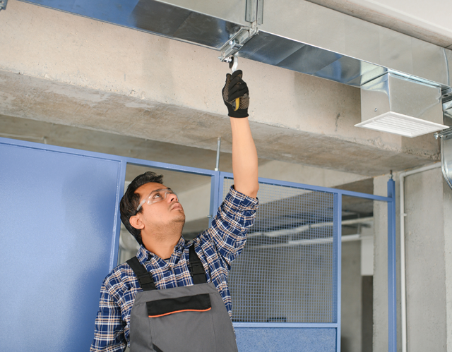 Man in work clothes inspecting ductwork, reaching up with a tool. Industrial setting.