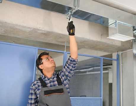 Man in work clothes inspecting ductwork with a tool. Indoors, concrete ceiling, blue door, and metal cage in background.