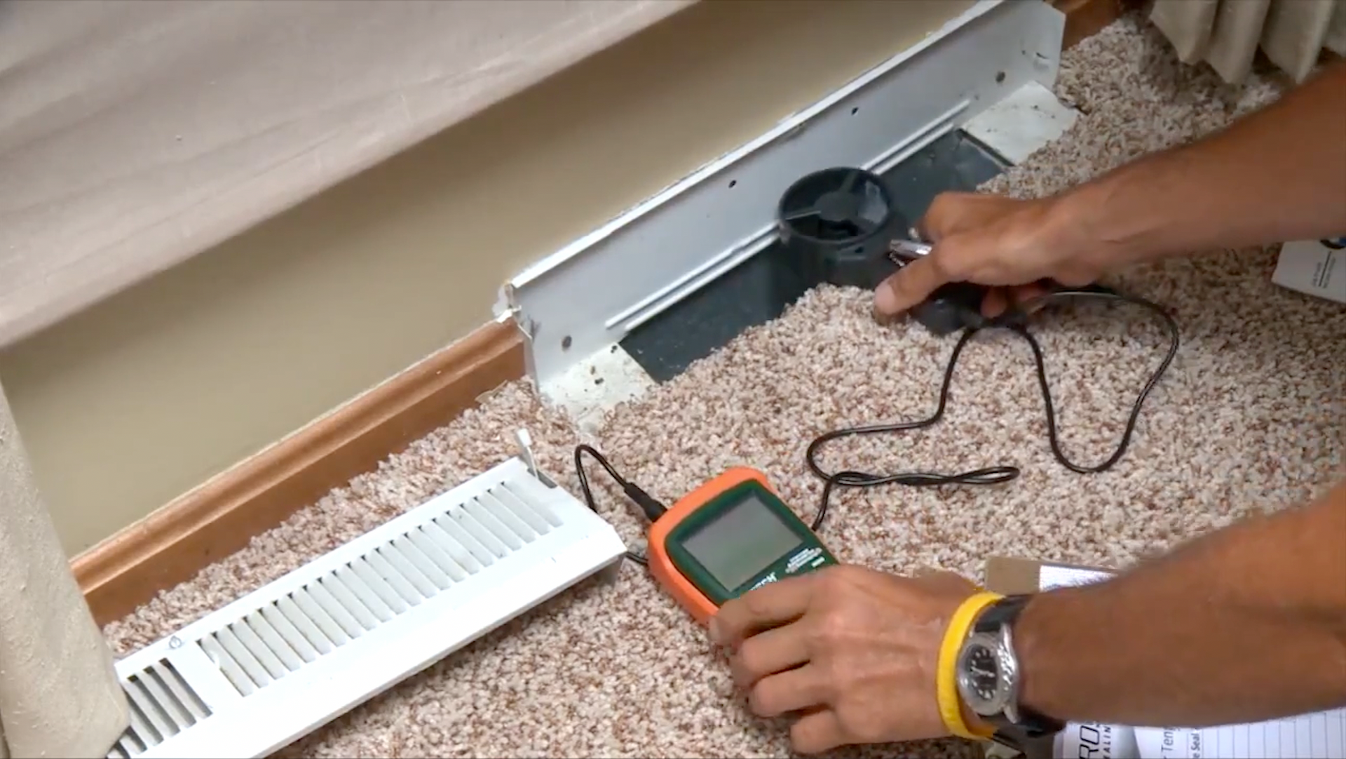 Person testing air vent with a handheld meter on carpeted floor near wall.