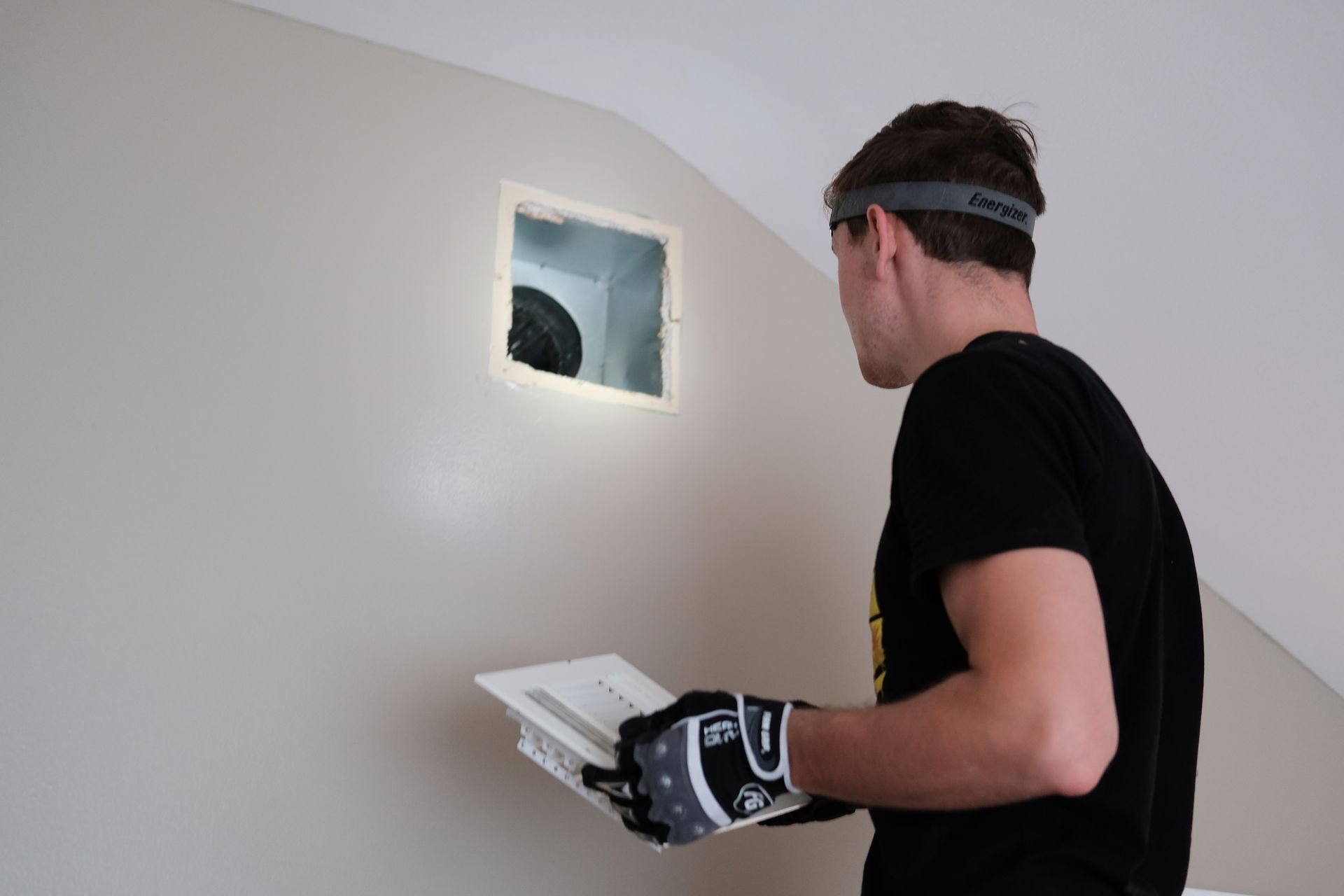 Person looking at square hole in wall; holding vent cover; wearing work gloves and headband.