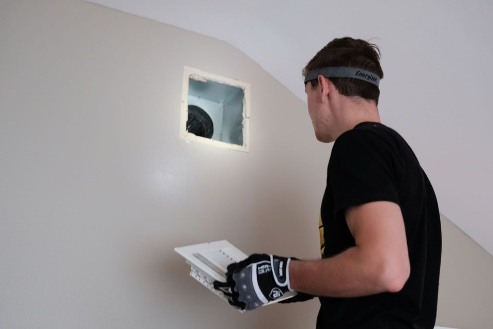 Person with headlamp and gloves, holding vent cover, looking at recessed opening in wall.