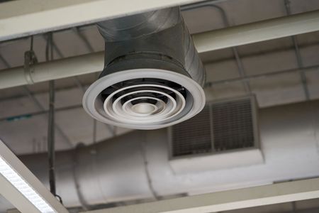 Ceiling air vent with concentric circular baffles, near a rectangular vent, gray ducts, and white ceiling beams.