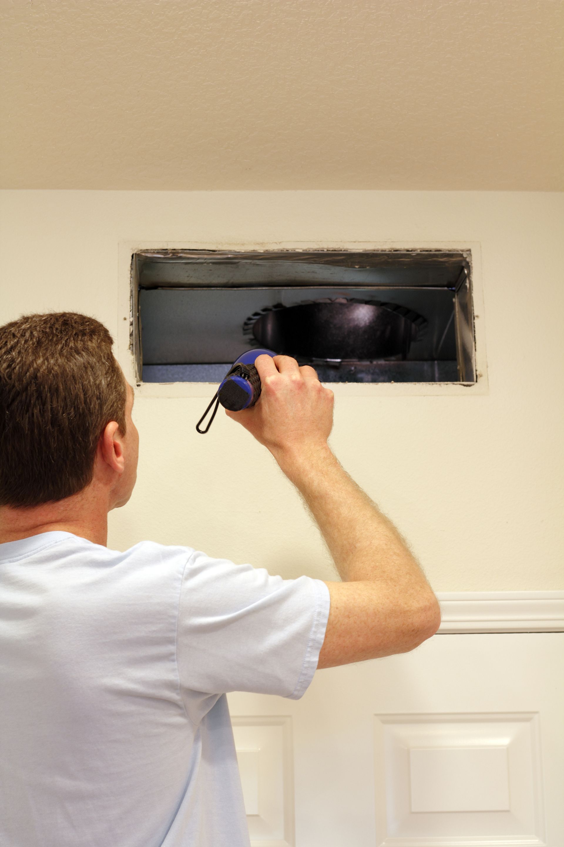 Man using a flashlight to inspect an open vent in a white wall.