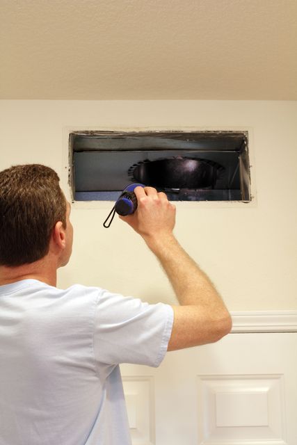 Man inspecting ductwork with a flashlight, looking into a rectangular opening in a white wall.