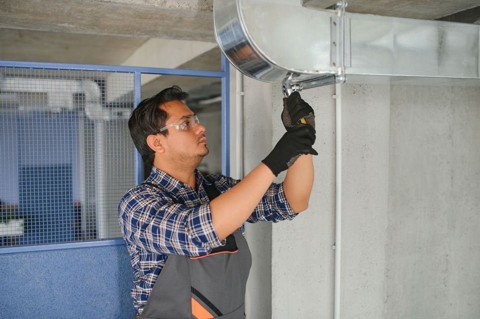HVAC technician installs ductwork. Wearing safety glasses and gloves, he works indoors near a wall.