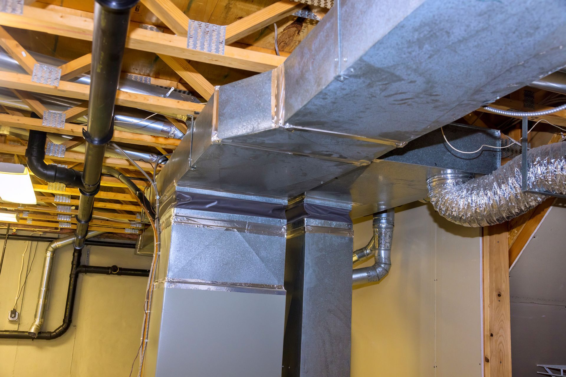 Ductwork and pipes in a basement. Silver metal ducts with insulation, black pipes, and exposed wooden beams.