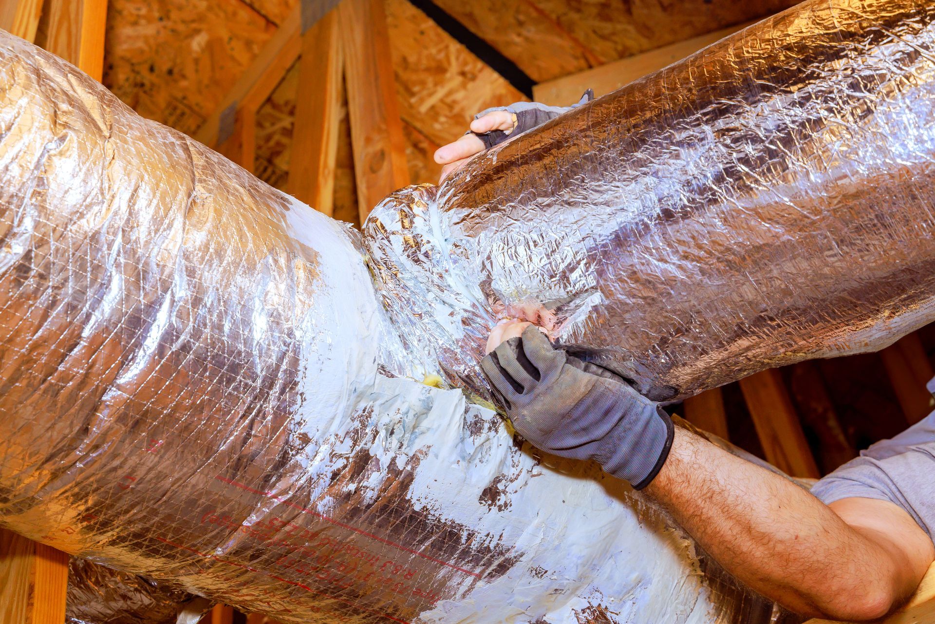 Person installing insulated ductwork in an attic, using gloved hands and sealant.