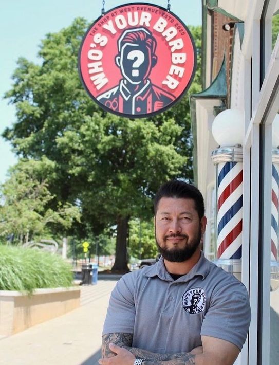 A man stands in front of a who 's your barber sign