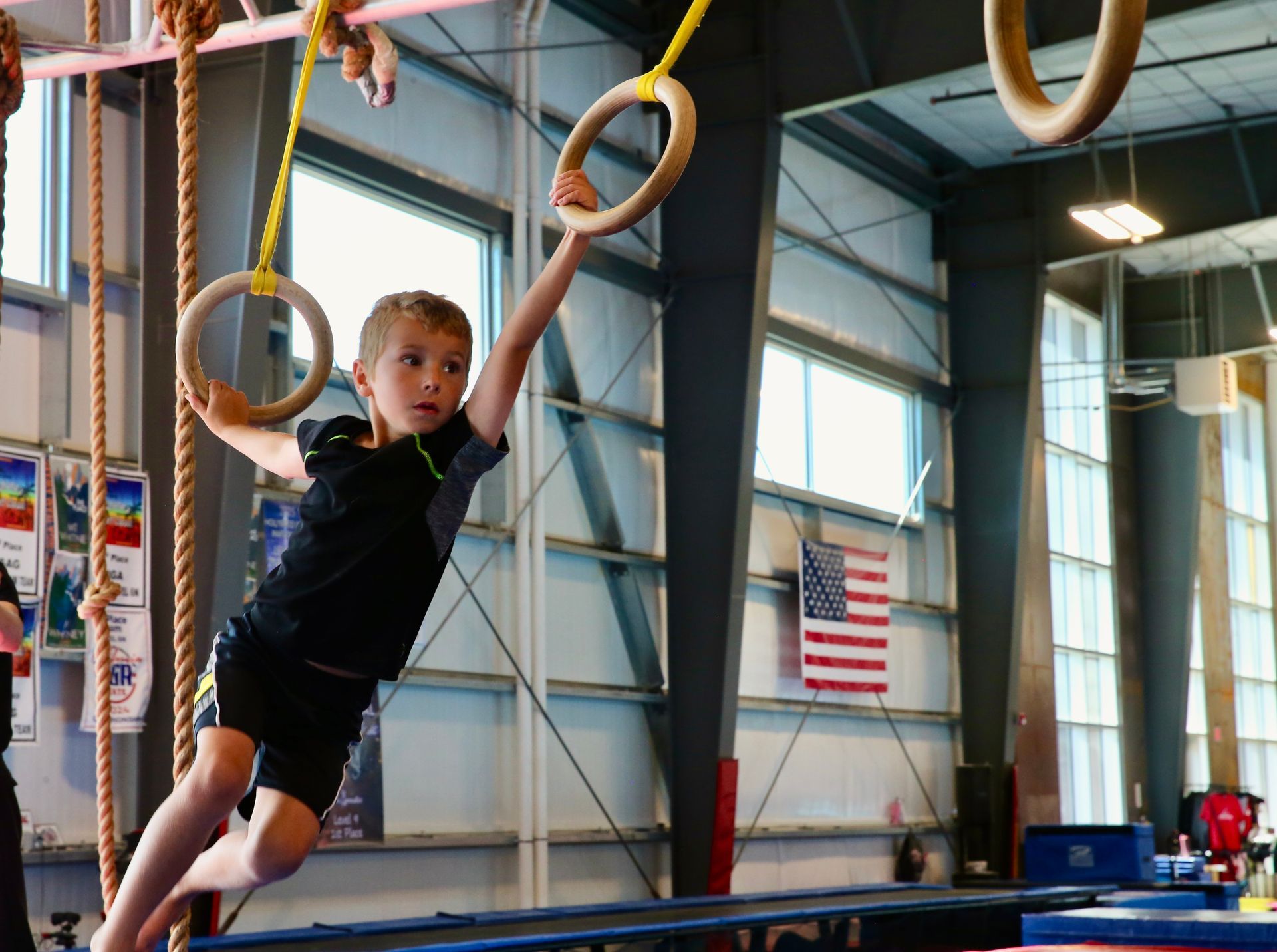 Child swinging on rings in an indoor gym, with an American flag and equipment in the background