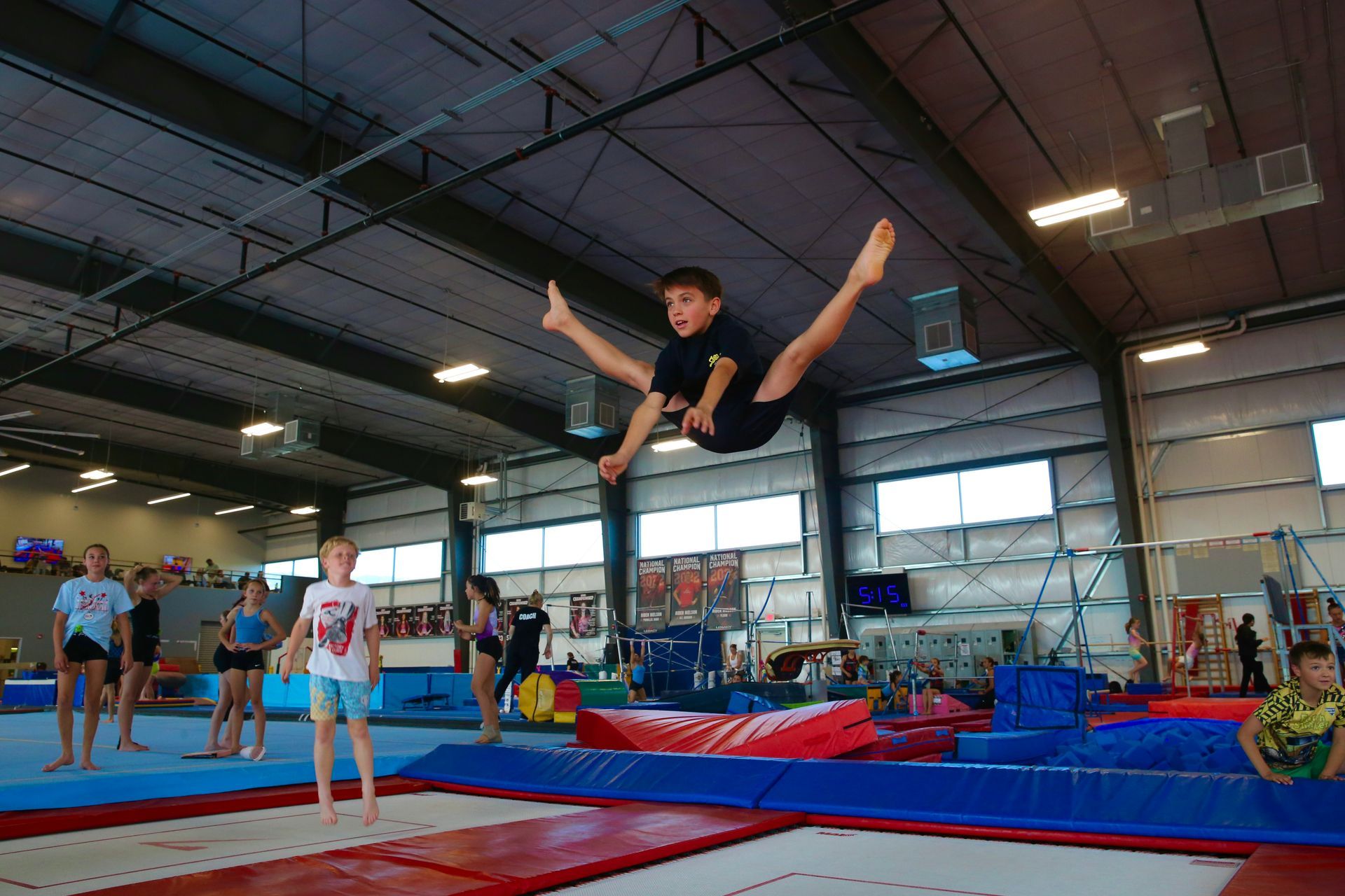 Gymnast mid-air in a split leap over blue mats inside a gymnasium, with other athletes nearby