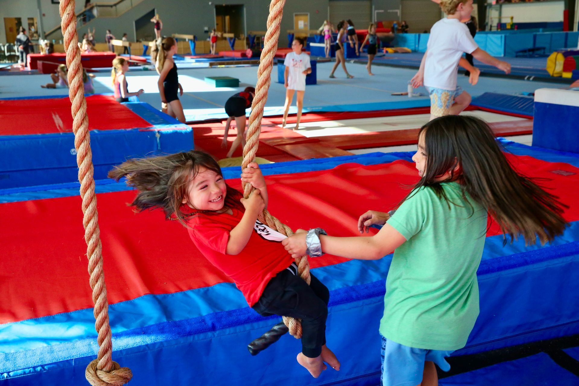 Children swinging on ropes over a red-and-blue padded gym floor, with other kids in the background