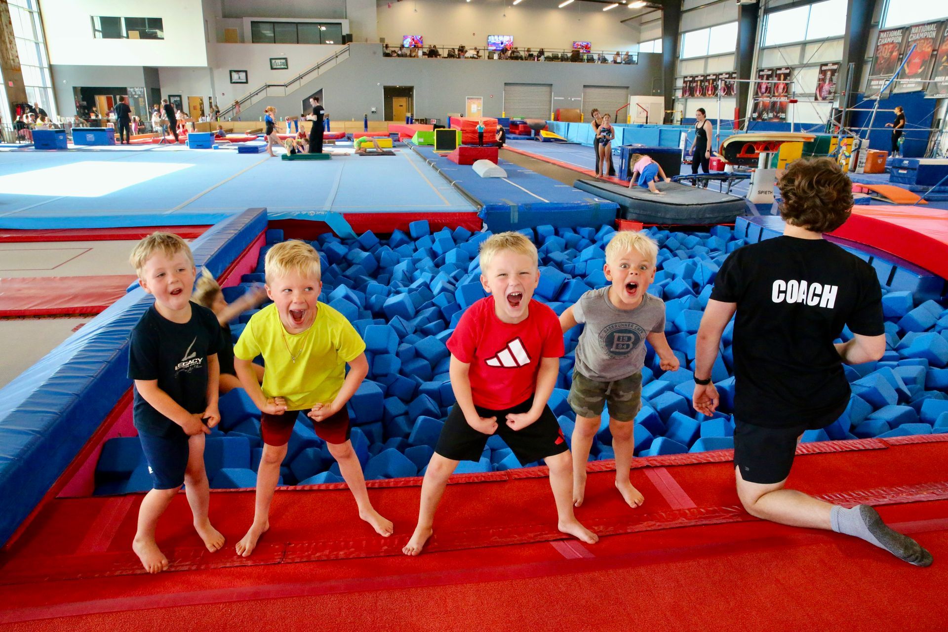 Children bouncing on a red mat in an indoor gymnastics gym, with a coach kneeling nearby.
