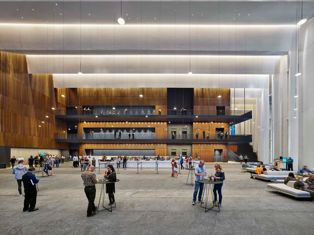 Large open hall with people mingling. A multi-level wooden structure is in the background. Gray floor, white walls.