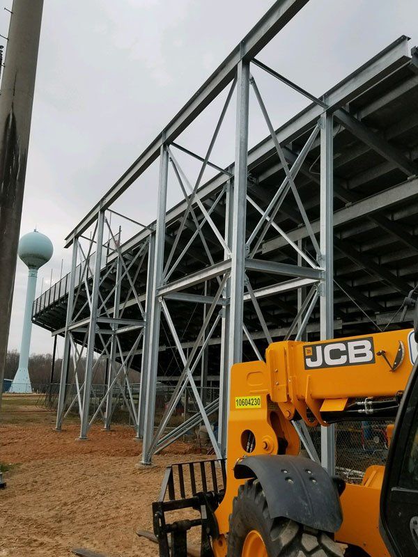 Steel bleachers being constructed with a JCB forklift in the foreground and a water tower in the background.