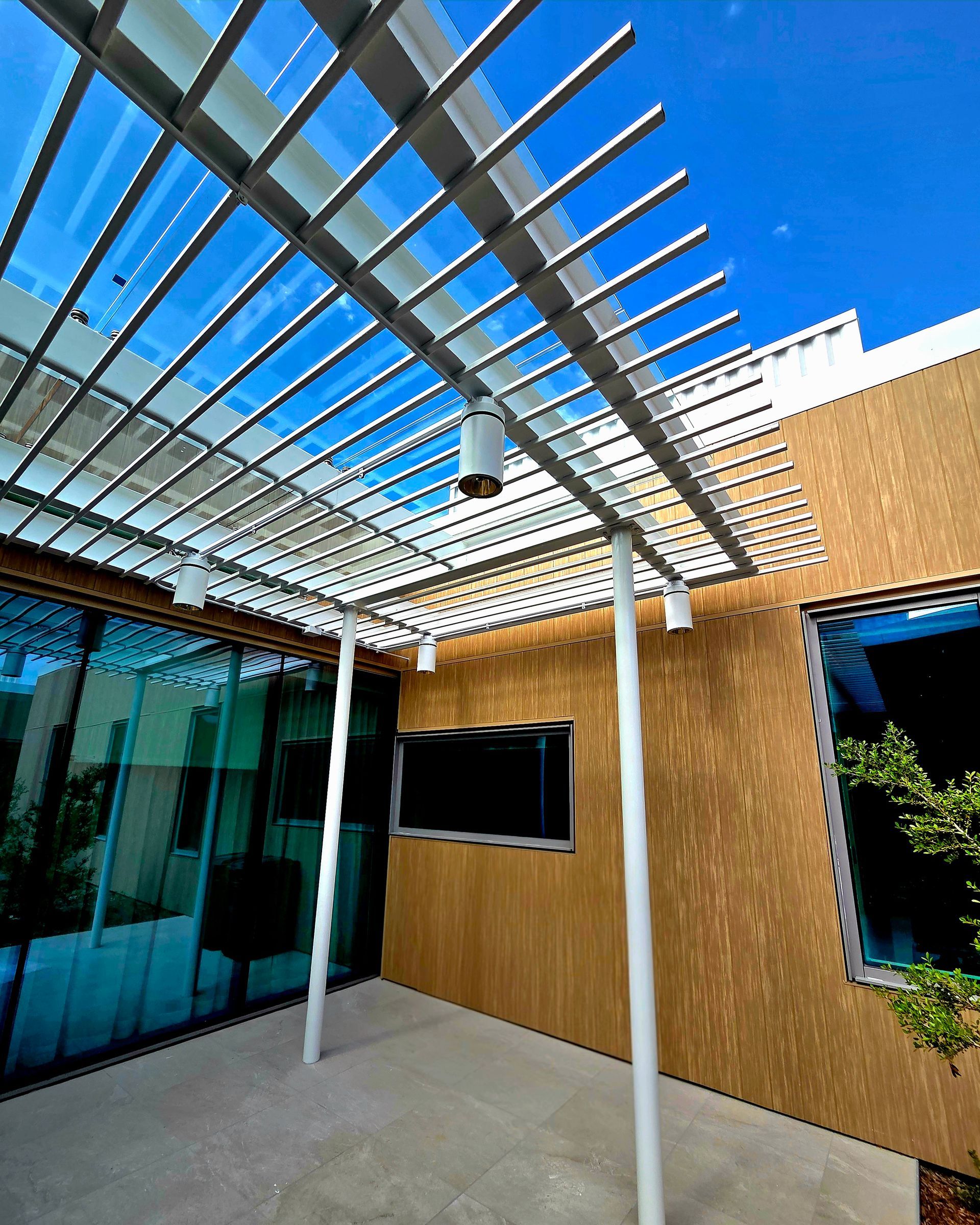 Outdoor patio with metal pergola, lights, and a mounted TV against a textured wood wall, blue sky.